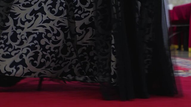 Close-up of a woman in a black and white dress with intricate patterns walking on the red carpet. The woman's legs in black shoes are visible from the hem of the dress. Side view