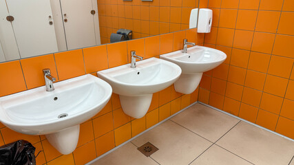 Modern public restroom with three white sinks against an orange tiled wall, hygiene, cleanliness