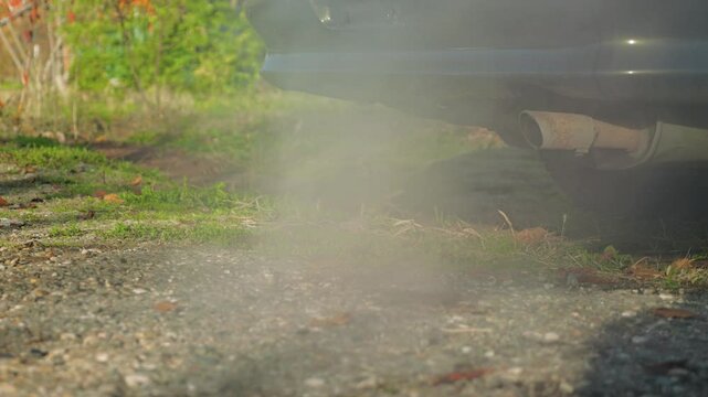 Exhaust pipe of a car parked on a dirt road and emitting noxious smoke into the grassy and wooded surroundings. The exhaust pipe is a little rusty. Slow motion