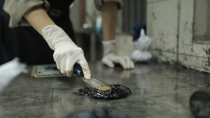 A close-up of A printmaker wearing gloves carefully spreads thick black ink on a metal intaglio...