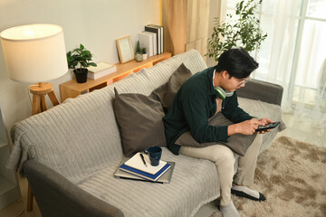 Modern Lifestyle, Balance Between Work and Relaxation, A young man in casual attire sits on a cozy couch, using his smartphone with headphones around his neck, taking a break at home