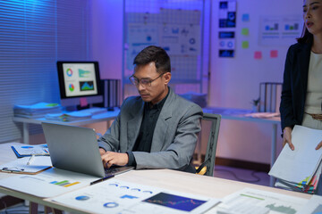 Professional working on a laptop, surrounded by charts and graphs in a modern office. Engaging in data analysis and business strategy planning, illuminated by blue ambient lighting