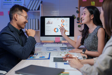 Asian businesspeople are discussing marketing strategy analyzing charts on computer screen showing financial data in office meeting at night