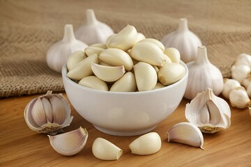 A white bowl filled with peeled garlic cloves, surrounded by whole garlic bulbs and scattered cloves on a wooden surface.