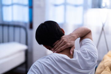 Asian man sitting on a bed holding his neck in discomfort, experiencing office syndrome pain or stiffness, possibly due to poor sleep or posture in a bedroom apartment