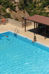 Elevated shot of a resort pool in the mountains with a girl in a bikini with a hat sitting on the curb and a boy floating in the water.