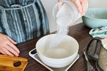 Woman cook in apron pouring sugar into bowl on kitchen scales on kitchen table