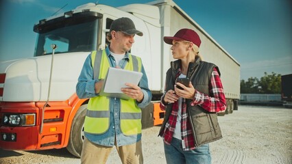 Caucasian woman and male standing next to each other. Man listening to female trucker talking after finishing delivery to requested location. Filling in data on notebook. Truck in background.