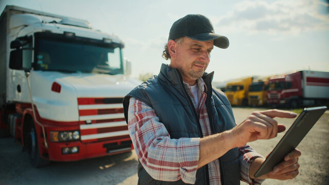 Handsome Caucasian trucker standing in front of long truck. Using tablet device while chatting with customer. Looking at camera and smiling with joy. Wearing baseball cap and warm vest.