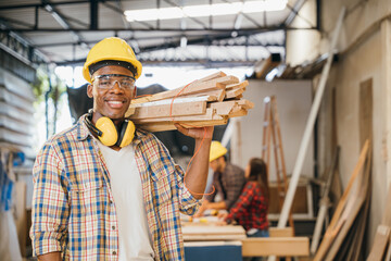 Smiling construction worker in a yellow hardhat and plaid shirt holding wood planks on his...