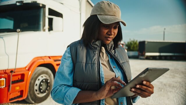 Charming African American woman with cap and blue shirt standing in front of her truck. Working in distribution company. Using tablet device for confirming completed order to location.