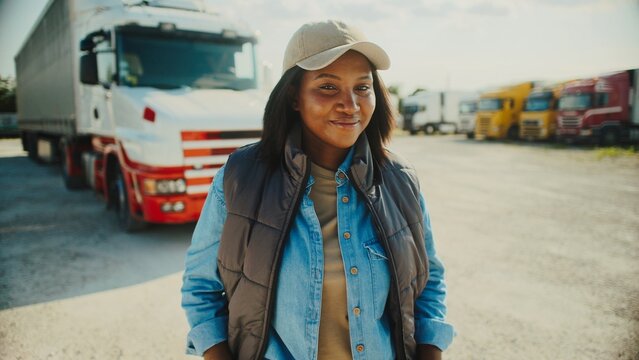 Cute African American woman standing in front of truck parking lot. Looking at camera while smiling. Resting in fresh air. Clear sky with sun shining bright. Morning before trucker departs to location - Powered by Adobe