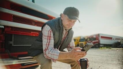 Positive Caucasian man wearing warm vest and baseball cap. Sitting on bumper of long lorry. Looking...