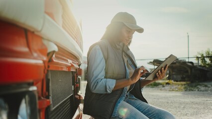 Naklejka premium Side camera view of focused African American female sitting on drop off bench at front of her truck. Driver looking at tablet screen. Typing with fingers on display. Sun shining bright in background.