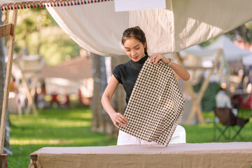 Young asian saleswoman showcasing a checked scarf at her outdoor market stall, enthusiastically promoting her textile products to attract potential customers amidst a vibrant setting