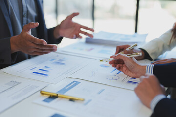 Business professionals engaged in a collaborative meeting, analyzing financial documents and charts. Hands point to graphs and data on papers, indicating a focused discussion on strategy
