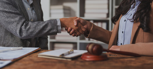 Two business professionals shaking hands across a desk in an office. A gavel and documents are visible, suggesting a legal or corporate agreement taking place