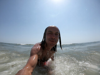 Handsome guy with long hair having fun at beach, sea waves splashing on his body