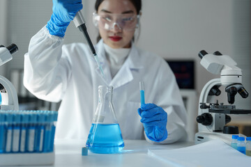 Scientist in a lab coat and gloves dropping blue liquid from a pipette into an erlenmeyer flask,...
