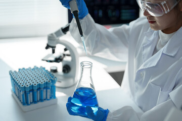 Scientist wearing lab coat, gloves and glasses using micropipette to drop blue reagent into...