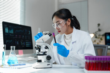 Young female scientist wearing lab coat and gloves carefully examining blood sample near microscope in modern laboratory, with computer displaying data analysis on screen
