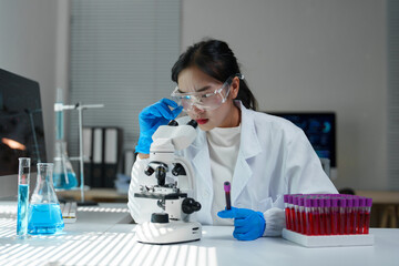 Young female scientist adjusting microscope while holding blood sample tube, conducting medical...