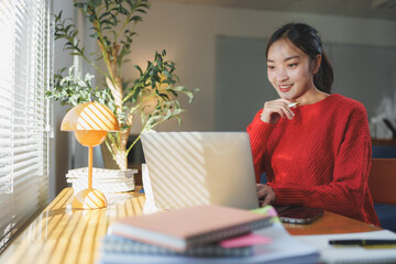 Young woman in a cozy setting works remotely on her laptop. Natural light filters through blinds,...