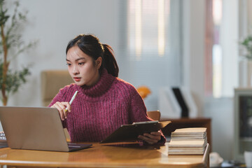 Focused young woman in a cozy home setting, analyzing data on a laptop. Holding a clipboard and pen, surrounded by books, suggesting a study or remote work environment