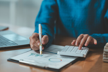 Person in blue sweater analyzing financial data using a calculator and laptop, focusing on charts and graphs on a clipboard, highlighting business and finance concepts