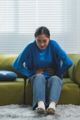 Young woman in a blue sweater sitting on a green couch, holding her stomach in discomfort, possibly experiencing abdominal pain or cramps, with a concerned expression