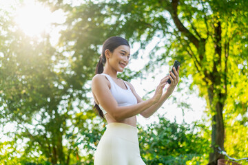 Obraz premium Smiling Asian woman using smartphone in green park after workout. Young active woman in fitness wear using phone while enjoying healthy lifestyle outdoors.