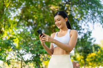 Asian woman using phone in park after workout. Young woman in sportswear smiling while checking smartphone outdoors in nature.