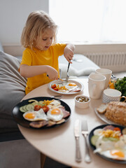 Hungry child curly blonde girl happily eats food with fork and knife on her own. vertical.
