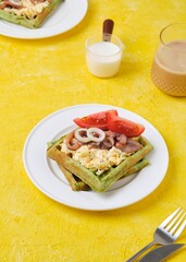 Breakfast, spinach waffles with egg salad, pickled herring and fresh tomato on a white plate on a yellow concrete background. Waffle recipes.