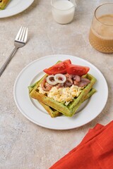Breakfast, spinach waffles with egg salad, pickled herring and fresh tomato on a white plate on a light concrete background. Waffle recipes.
