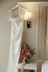 An elegant wedding dress was hanging up and a bouquet of multicolored flowers was next to it on the bedside table