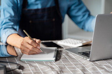 Entrepreneur taking notes while planning an online startup business at a desk with a laptop and...