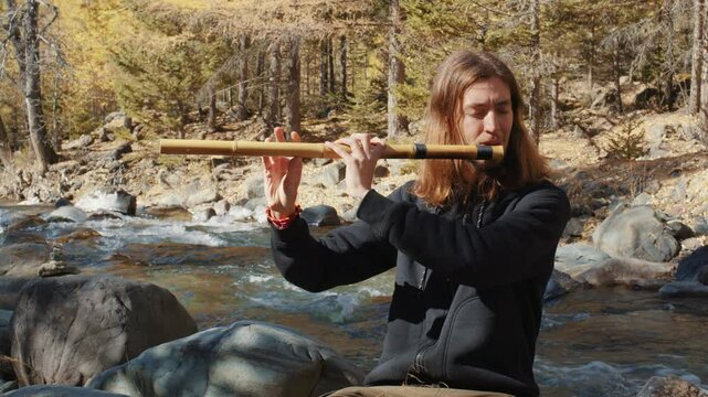 Young musician playing wooden flute while sitting near flowing river, surrounded by golden autumn forest with vibrant fall colors