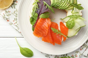 Delicious bagel with salmon, cream cheese, cucumber and avocado on white wooden table, flat lay
