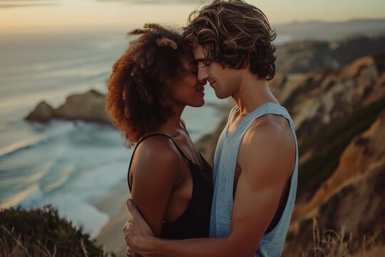 Authentic interracial couple dressed casually, kissing affectionately while facing one and other and holding hands with ocean and mountains in the view behind them, - Powered by Adobe