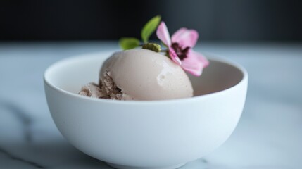 White bowl filled with a scoop of ice cream. the ice cream is light brown in color and has a smooth texture. on top of the ice cream, there is a small pink flower with green leaves.