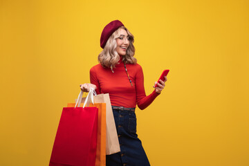 Stylish woman in bright outfit holding shopping bags and interacting with her phone, reflecting the joy of shopping.