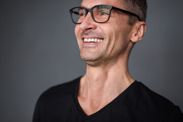 A cheerful man wearing glasses and a dark shirt, smiling and looking relaxed against a simple background. His expression radiates positivity and approachability.
