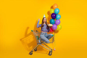 Cheerful young woman celebrating with balloons and cake in a shopping cart against yellow background