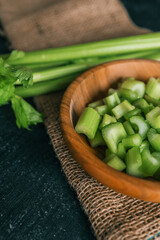 Fresh celery stalks and chopped pieces in a wooden bowl on a rustic dark table. Organic green vegetable on burlap with wooden background. Healthy eating, nutrition, and cooking ingredients concept