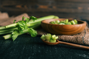 Fresh chopped celery in a wooden bowl with whole celery stalks on a rustic dark surface. Healthy organic food concept with natural textures and earthy tones