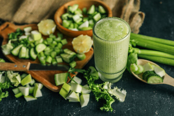 Fresh green smoothie in a textured glass surrounded by chopped ingredients including cucumber,...
