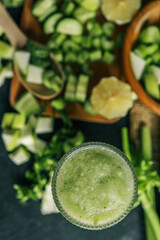 Fresh green smoothie in a textured glass surrounded by chopped ingredients including cucumber,...