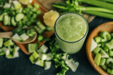 Fresh green smoothie in a textured glass surrounded by chopped ingredients including cucumber,...