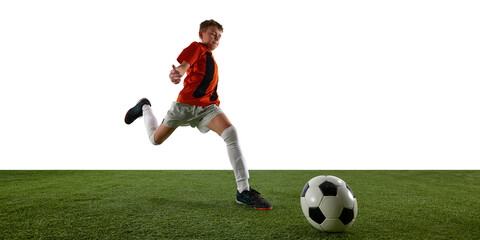 Dynamic image of focused boy, soccer player in orange jersey in motion on field, running towards ball against white background. Concept of sport, education, competition, game